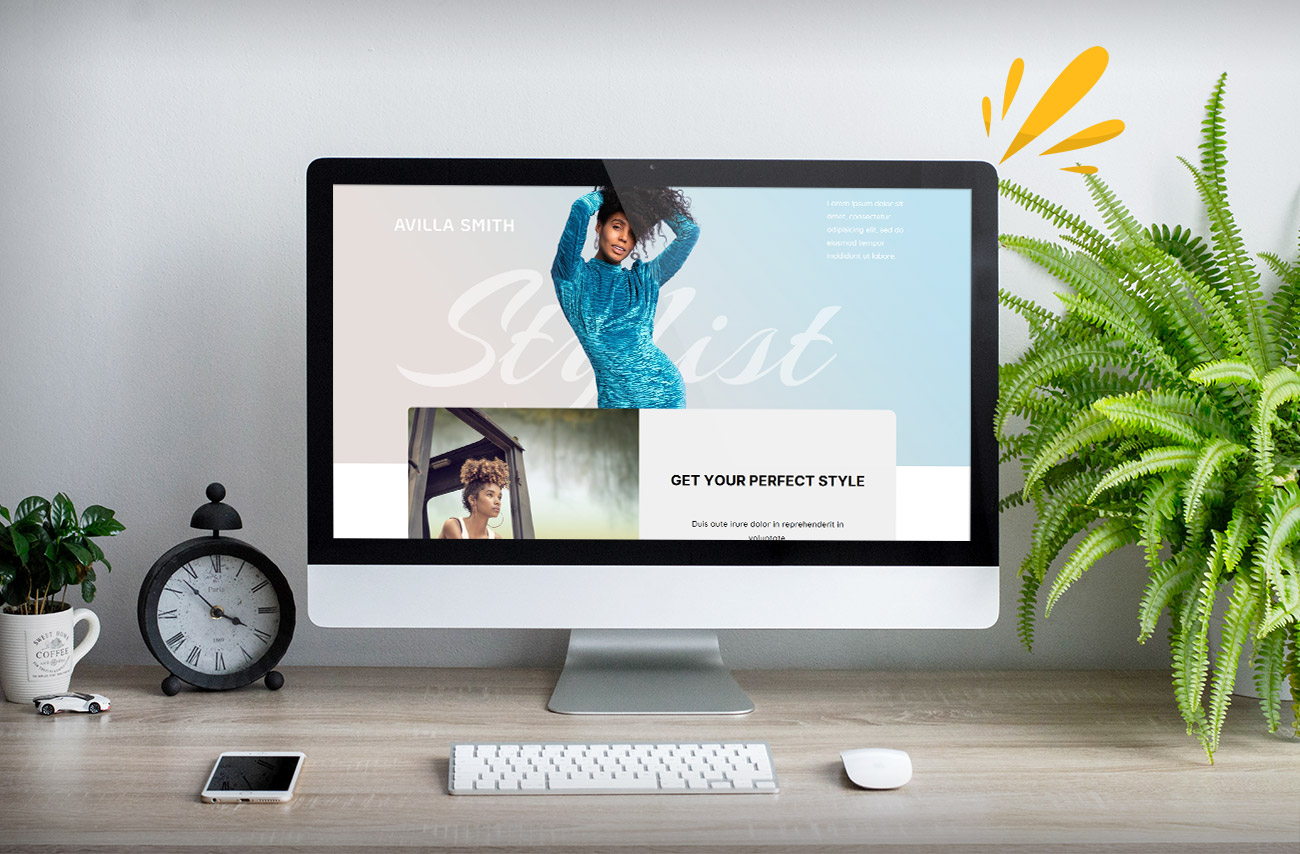 A computer display shows a stylist's website featuring a blue dress, photos of clients, and a plant on a wooden desk with a clock.